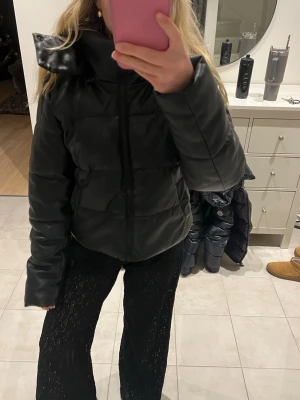 Svart pufferjacka i skinn - Säljer en svart pufferjacka i skinnimitation med dragkedja framtill och luva. Jackan har en oversized känsla och är perfekt för kyliga dagar. Den har långa ärmar och en stilren look som passar till många outfits. Den är superskön och håller än varm på vintern, knappt använd bara testad. 