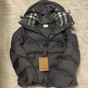 Svart pufferjacka från Burberry - Säljer en svart pufferjacka från Burberry med avtagbara ärmar så du kan bära den som väst. Jackan har huva med klassiskt rutigt foder, dragkedja och knappar framtill samt tydlig Burberry-patch på ärmen. Perfekt för kalla dagar och riktigt snygg streetstil.