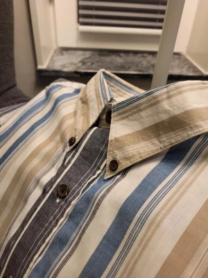 Randig skjorta från Adelsten L - Snygg kortärmad skjorta från Adelsten Clothing Company i storlek L. Skjortan har breda ränder i beige, blått, vitt och grått, med bruna knappar och två bröstfickor. Materialet är mjuk bomull, perfekt för chill dagar. Klassisk krage med knappdetaljer.