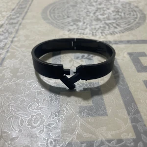 Svart armband Hermer - Stilrent svart armband med cool H-formad låsdetalj framtill. Armbandet har en modern och minimalistisk design med slät yta och är tillverkat i ett hårt material som ger en edgy känsla. Perfekt för dig som gillar att sticka ut med accessoarer som har en unik twist.
