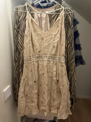 Beige kortklänning med paljetter - Supersöt beige kortklänning från mallorca/spanien. med glittriga paljettdetaljer och broderad midja. Klänningen har v-ringad hals, flätade snören med tofsar och ett skirt, luftigt material som känns som bomull. Perfekt för sommarens alla tillfällen och ger en boho-vibe. Den är verkligen jätte fin och har kommit till användning så många somrar. Dock har en tofs på snöret försvunnit och vid urringningen på klänningen har de gått sönder lite men det går att sy lätt. Osäker på strlk men ungefär XS/S