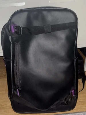 Svart DB ryggsäck med lila detaljer - Säljer en stilren svart ryggsäck med lila detaljer och svarta remmar. Väskan har en rektangulär form, dragkedja framtill och justerbara axelremmar. Perfekt för dig som vill ha en enkel och snygg väska till skolan eller utflykten.