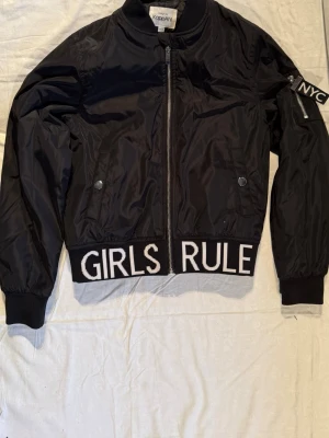 Svart bomberjacka från KappAhl - Svart bomberjacka från KappAhl med coola detaljer som texten 'GIRLS RULE' längs nederkanten och en NYC-patch på ärmen. Jackan har dragkedja framtill, ribbade muddar och fickor med tryckknappar. Perfekt för dig som vill sticka ut.
