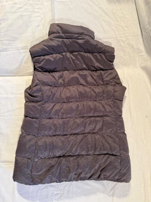 Grå dunväst från Uniqlo, storlek S - Säljer en grå dunväst från Uniqlo i storlek S. Västen har quiltad design och dragkedja framtill. Perfekt för lager-på-lager och har en hög krage som skyddar mot vind. Materialet är mjukt och lätt, vilket gör den enkel att bära under höst och vinter.