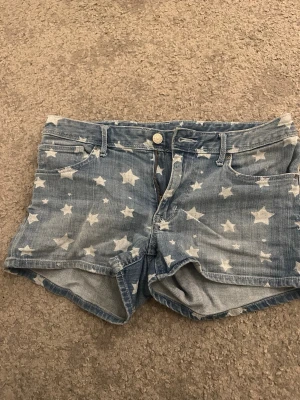 Blå jeansshorts med stjärnor från H&M - Säljer ett par blå jeansshorts från H&M med coolt stjärnmönster över hela tyget. Shortsen har klassisk femficksdesign, knapp och dragkedja framtill samt bälteshällor. Perfekta för sommaren och har en loose passform.