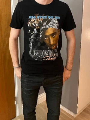 Svart 2Pac t-shirt All Eyez On Me - Svart t-shirt med tryck av 2Pac och texten 'All Eyez On Me' i blått. T-shirten har klassisk passform, rund hals och korta ärmar. Materialet känns mjukt och skönt, perfekt för dig som gillar streetwear och hiphop-vibe.