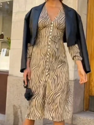 Zebramönstrad långklänning från Adoore - Snygg långklänning från Adoore med zebramönster i beige och mörkbrun. Klänningen har v-ringning, långa ärmar med resår vid ärmslut och dekorativa knappar framtill. Tillverkad i ett lätt, transparent syntetmaterial som ger ett luftigt intryck.