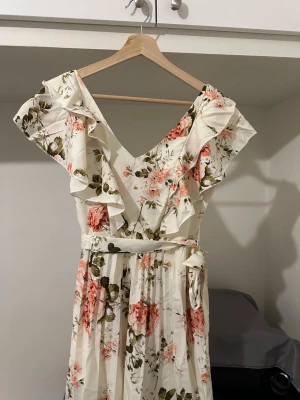 Blommig kortklänning med volang - Supersöt kortklänning från Elenzga med blommigt mönster i rosa och grönt på vit botten. Klänningen har v-ringning, volangdetaljer vid ärmarna, markerad midja med knytband och lätt rynkad kjol. Perfekt för sommarens alla tillfällen!