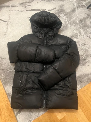 Svart pufferjacka från FSBN XS-S - Svart pufferjacka från FSBN i storlek XS  Jackan har en stor huva, breda kanaler och cool dragkedja med CHAOTIC-anhängare. Perfekt för kalla dagar och har två sidofickor. Materialet är syntet och jackan är långärmad.