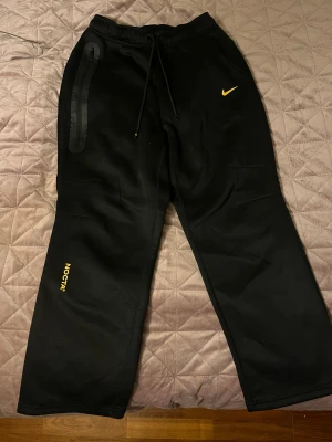 Nike NOCTA svarta mjukisbyxor - Svarta mjukisbyxor från Nike NOCTA med gul Swoosh på framsidan och broderade detaljer som NOCTA, A.W.R. och C.L. på benen. Byxorna har snörning i midjan, dragkedja på ena fickan och är tillverkade i mjukt bomullsmaterial. Snygg loose passform och stilren look.