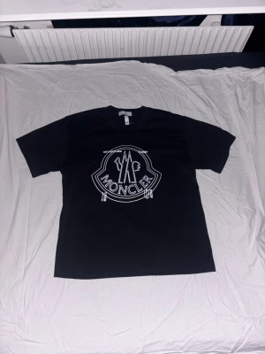 Svart Moncler t-shirt med stor logga - Kom med pris. Svart t-shirt från Moncler med stor vit logga tryckt på bröstet. Klassisk rund hals och korta ärmar. Materialet är mjuk bomull som känns skönt mot huden. Perfekt för dig som gillar streetwear och vill ha en clean look med tydlig branding.