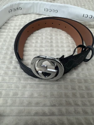 Svart G bälte  - Snyggt svart bälte med mönster och ett stort, rundat spänne i silverfärgad metall. Bältet är gjort i skinn och har en stilren design som lyfter vilken outfit som helst. Insidan är brun och bältet kommer med originalpåse.