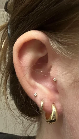 Guldpläterade örhängen från Montini - Säljer ett par chunky, runda örhängen i guldpläterad metall från Montini. De har en stilren, blank finish och är perfekta för att ge din look lite extra edge. Passar dig som gillar klassiska hoops med modern twist.