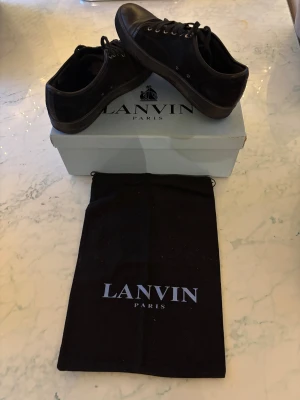 Svarta sneakers från Lanvin Paris - Snygga svarta sneakers från Lanvin Paris med rund tå och platt sula. Ovandelen är i mocka med detaljer i skinn på tå och häl. Skorna har snörning med metallöglor och kommer med originalkartong och dustbag. Perfekta för dig som gillar stilrena och exklusiva sneakers.