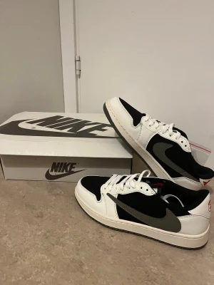 Nike Jordan 1 Low svart/vit/grå - Nike Jordan 1 Low sneakers med svart och vit ovandel i skinn och textil, grå swoosh och matchande grå yttersula. Klassisk rund tå, snörning och platt sula. Snygg och stilren färgkombo som passar till streetwear och sneakers-samlingen.