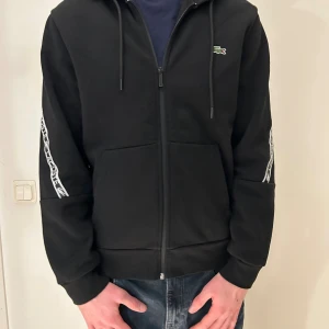 Svart hoodie från Lacoste med logoband - Svart hoodie från Lacoste med dragkedja och huva. Snygga vita logoband längs ärmarna och klassisk krokodillogo på bröstet. Tröjan har snörning vid huvan och två fickor framtill. Perfekt för chill eller streetwear.
