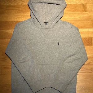 Grå hoodie från Polo Ralph Lauren - Snygg grå hoodie från Polo Ralph Lauren med klassisk broderad logga på bröstet. Tröjan har huva, långa ärmar och en mjuk bomullskänsla. Perfekt för chill dagar och enkel att matcha med jeans eller joggers. Inga synliga tecken på att den har vart använd, använt ett fåtal gånger. Ordinarie pris 1200kr. Säljs för endast 499kr. Pris diskuterbart. 