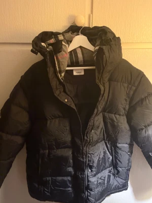 Burberry Vinterjacka  - Svart pufferjacka från Burberry med klassisk rutig huva i grått, vitt och rött. Jackan har dragkedja och tryckknappar framtill, samt en varm och fluffig känsla. Perfekt för kalla dagar och har en snygg, modern look med märkets ikoniska detaljer. Ej Äkta!