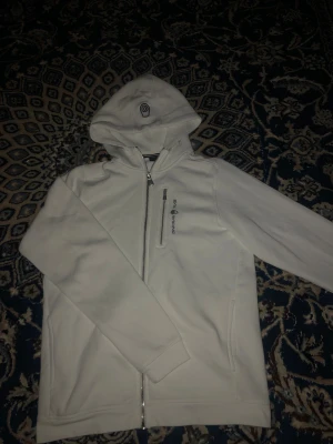 Vit hoodie från Sail Racing med huva - Vit hoodie från Sail Racing med broderad logga på huvan och bröstet. Tröjan har hel dragkedja framtill, en extra bröstficka med dragkedja och två sidofickor. Mjuk insida och klassisk passform, perfekt för chill dagar. Storlek 176=S