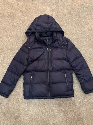 Mörkblå pufferjacka Polo Ralph Lauren  - Snygg mörkblå pufferjacka från Polo Ralph Lauren i storlek XL (Kidsbrandstore). Jackan har huva, flera dragkedjor och den klassiska röda Polo-loggan på bröstet. Fylld med dun och har två sidofickor med dragkedja. Perfekt för kalla dagar. Nypris: 3000 kr på Kidsbrandstore. Därmed är storleken XL i barnstorlek vilket motsvarar S i herr storlek, kan användas för tjejer också 