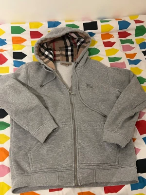 Grå Burberry hoodie med dragkedja - Säljer en grå hoodie från Burberry med klassiskt rutigt foder i huvan och insidan. Tröjan har dragkedja, snörning vid huvan och ribbade muddar. Ikonisk Burberry-logga broderad på bröstet. Perfekt för dig som gillar stilrena och lyxiga streetwear-plagg.