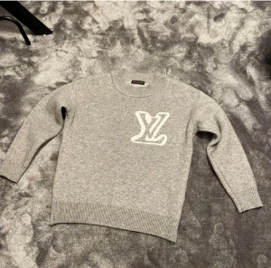 Grå stickad Louis Vuitton tröja - Snygg grå stickad tröja från Louis Vuitton med stort vitt LV-logo framtill. Tröjan har rund hals, ribbade muddar och långärmad passform. Perfekt för dig som gillar exklusiva märken och vill ha en stilren look.
