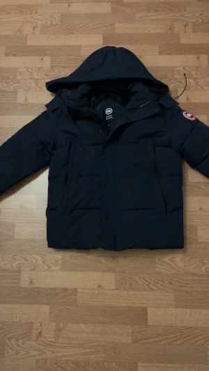 Svart dunjacka från Canada Goose S - Säljer en svart dunjacka från Canada Goose med huva och klassisk logga på ärmen. Jackan har dragkedja och knappar framtill samt stora fickor. Perfekt för kalla dagar och riktigt snygg streetstil.