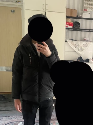 Svart Sail Racing pufferjacka med huva - Svart pufferjacka från Sail Racing, modell Antarctica Expedition. Jackan har huva, två bröstfickor med dragkedja och snygga logodetaljer på bröstet och ärmen. Perfekt för kalla dagar med sin tjocka och isolerande design. Stängs med dragkedja framtill.