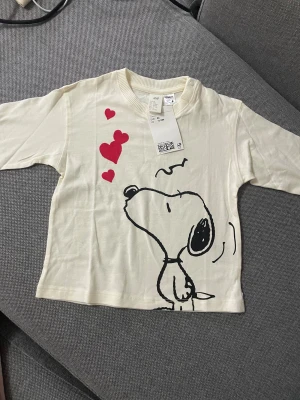 Vit långärmad Snoopy-tröja H&M - Vit långärmad tröja från H&M med stort tryck av Snoopy och röda hjärtan på framsidan. Rund hals med ribbad kant och tryckknappar vid axeln för enkel påklädning. Mjuk och skön bomull som passar perfekt för chill dagar.