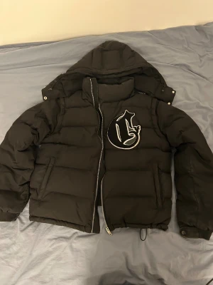 Svart pufferjacka från Corvidae M - Svart pufferjacka från Corvidae med avtagbar huva och stor broderad logga på bröstet. Jackan har dragkedja framtill, två sidofickor och justerbar nederkant med snörning. Perfekt för kalla dagar och har en clean, urban vibe.