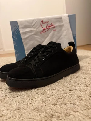 Christian Louboutin svarta sneakers mocka - Svarta sneakers från Christian Louboutin i lyxig mocka med klassisk röd sula. Skorna har rund tå, snörning och diskret broderad logga på hälen. Insidan är ljusbeige och sulan är platt. Perfekta för dig som vill ha en clean och exklusiv look.