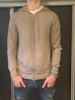 Beige cashmere hoodie från John Henric - Mjuk och lyxig beige hoodie i 100% cashmere från John Henric. Dragkedja framtill, två fickor och justerbar huva med snören. Perfekt för dig som gillar stilrena och bekväma plagg med premiumkänsla. Storlek M modellen på Biden är 182 väger 73 kg priset är ej stuket i sten! Hör av vid funderingar 