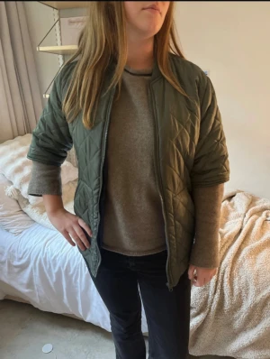 Militärgrön tre kvarts bomberjacka - Säljer en olivgrön bomberjacka med quiltat mönster och dragkedja framtill. Jackan har en klassisk bombermodell med ribbade muddar och är perfekt för dig som vill ha en stilren och trendig look. Passformen är normal och materialet känns lätt och smidigt.