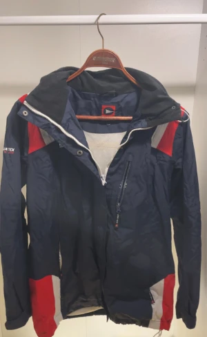 Marinblå vindjacka från Marine - Snygg marinblå vindjacka från Marine med röda och vita detaljer. Jackan har huva, meshfoder, dragkedja framtill och praktiska fickor. Perfekt för blåsiga dagar och har en sportig vibe med coola färgblock. Materialet är lätt och i syntet.