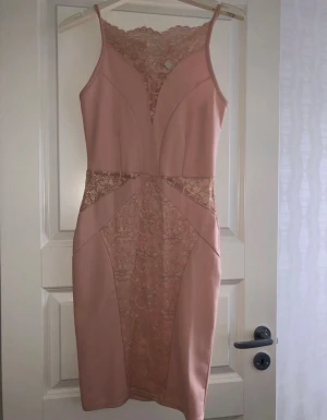 Rosa spetsklänning med smala axelband - Supersnygg rosa kortklänning med spetsdetaljer och smala axelband. Klänningen har figurnära passform, v-ringning och genombrutna spetspartier både fram och bak. Perfekt för dig som vill sticka ut med en feminin och trendig look.