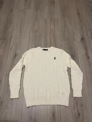Vit kabelstickad tröja från Polo Ralph Lauren - Säljer en klassisk vit kabelstickad tröja från Polo Ralph Lauren med broderad logga på bröstet. Tröjan har rund hals, ribbade muddar och långärmad passform. Perfekt för dig som gillar stilrena och tidlösa plagg.