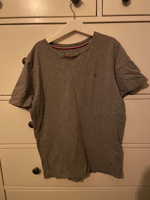 Grå t-shirt från Tommy Hilfiger - Snygg grå t-shirt från Tommy Hilfiger med liten broderad logga på bröstet. Klassisk rund halsringning och korta ärmar. Tillverkad i mjuk bomull för en skön känsla. Perfekt till jeans eller shorts.