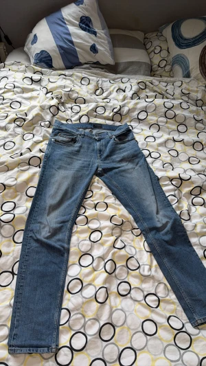 Tiger of Sweden Pistolero jeans blå - Snygga Tiger of Sweden Pistolero jeans i klassisk blå tvätt. Modellen har raka ben, fem fickor och detaljer med nitar. Tillverkade i Italien av slitstarkt jeanstyg med orange sömmar och knappstängning framtill. Perfekt för dig som gillar stilrena jeans.