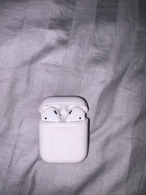 Apple AirPods 1 - Säljer ett par Apple AirPods 1 med laddningsetui. Hörlurarna är trådlösa och fungerar med både iPhone och andra enheter via Bluetooth. Etuiet har synligt slitage och repor, men hörlurarna ser ut att vara i gott skick. Svart skyddsfodral medföljer.