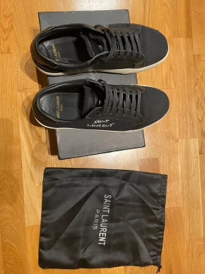 Svarta Saint Laurent sneakers - Svarta sneakers från Saint Laurent med vit broderad logga på sidan. Skorna har snörning, rund tå och platt vit sula. Ovandelen är i canvas med detaljer i skinn på hälen. Medföljer originalpåse och låda. Perfekt för dig som gillar stilrena designer.i skorna passar till 43 använda ett par gånger ‼️‼️först till kvarn som gäller ‼️