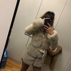 Beige pälsjacka med bälte - Säljer en beige pälsjacka med fluffiga detaljer på ärmar och kropp. Jackan har dragkedja framtill och ett matchande bälte i midjan för extra stilpoäng. Perfekt för dig som vill ha en mysig och snygg look under kalla dagar.