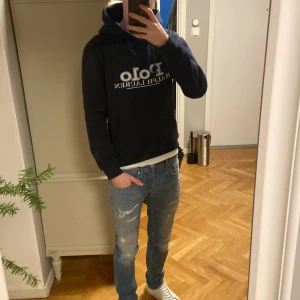 Mörkblå hoodie från Polo Ralph Lauren - Snygg mörkblå hoodie från Polo Ralph Lauren med stor vit logga på bröstet. Tröjan har huva med snörning, magficka och ribbade muddar vid ärmar och nederkant. Perfekt för dig som gillar klassisk streetwear och vill ha en bekväm och stilren look.