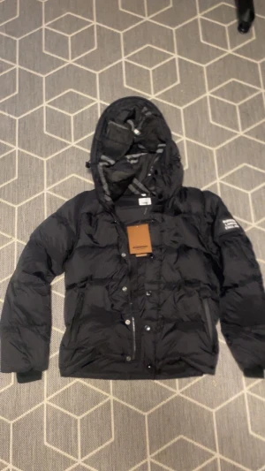 Svart Burberry pufferjacka med huva - Svart pufferjacka från Burberry med stor huva och klassiskt rutigt foder. Jackan har dragkedja och knappar framtill, samt en tydlig Burberry London England patch på ärmen. Perfekt för kalla dagar och har en clean, modern look.