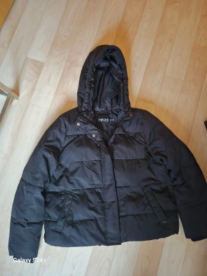 Svart pufferjacka från Pieces, M - Svart pufferjacka från Pieces i storlek M. Jackan har en stor huva, dragkedja framtill och två sidofickor. Den är quiltad och har en loose passform, perfekt för kalla dagar. Materialet är syntet och jackan är helsvart med en enkel, stilren look.