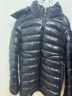 Svart Moncler pufferjacka med dragkedja - Säljer en svart pufferjacka från Moncler med glansig finish och klassisk logga på ärmen. Jackan har hög krage, två sidofickor med dragkedja och tydliga quiltade sektioner. Perfekt för dig som vill ha en snygg och varm jacka med exklusiv känsla.