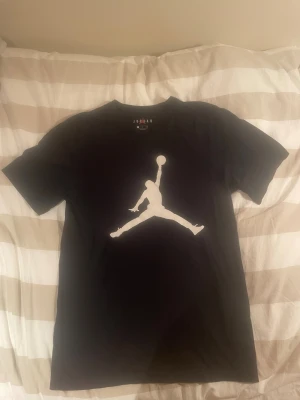 Svart Jordan t-shirt med logga - Säljer en svart t-shirt från Jordan med den ikoniska Jumpman-loggan i vitt på bröstet. T-shirten har rund hals och korta ärmar, perfekt för dig som gillar sportig och stilren streetwear. Materialet är mjuk bomull som känns skönt mot huden.