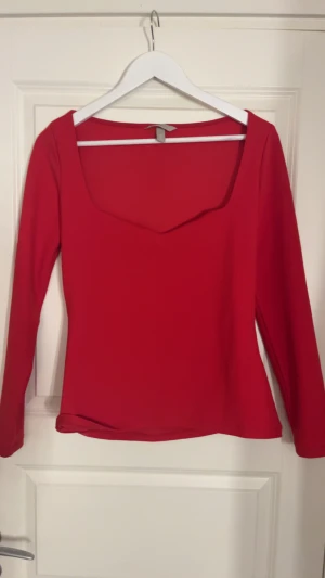 Röd långärmad topp med fyrkantig ringning - Snygg röd topp med lång ärm och en modern fyrkantig ringning. Toppens passform är normal och materialet känns mjukt och stretchigt, perfekt för dig som gillar stilrena och färgstarka plagg. Enkel design utan mönster eller extra detaljer.