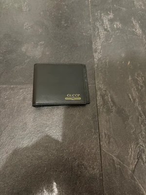 Svart plånbok från Gucci  - Snygg svart plånbok från Gucci i läder med guldfärgad logotyp på framsidan. Plånboken har flera kortfack och en stilren, klassisk design som passar perfekt i fickan. Perfekt för dig som gillar exklusiva accessoarer och vill ha något som sticker ut.