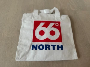 66 North vit väska - Vit shoppingväska från 66 North med stort rött och blått logotryck på framsidan. Väskan har två korta handtag och är tillverkad i slitstark syntet. Perfekt för att bära med sig grejer till skolan eller stan.