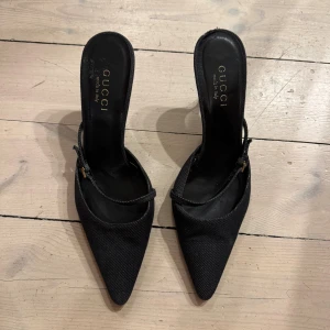 Svarta kitten heels från Gucci - Snygga svarta klackar från Gucci med spetsig tå och smal rem över foten. Skorna har elegant låg klack och är tillverkade i ett tygmaterial med diskret struktur. Guldiga detaljer på remmen ger en lyxig touch. Perfekta för dig som gillar stilrena och klassiska vibes.